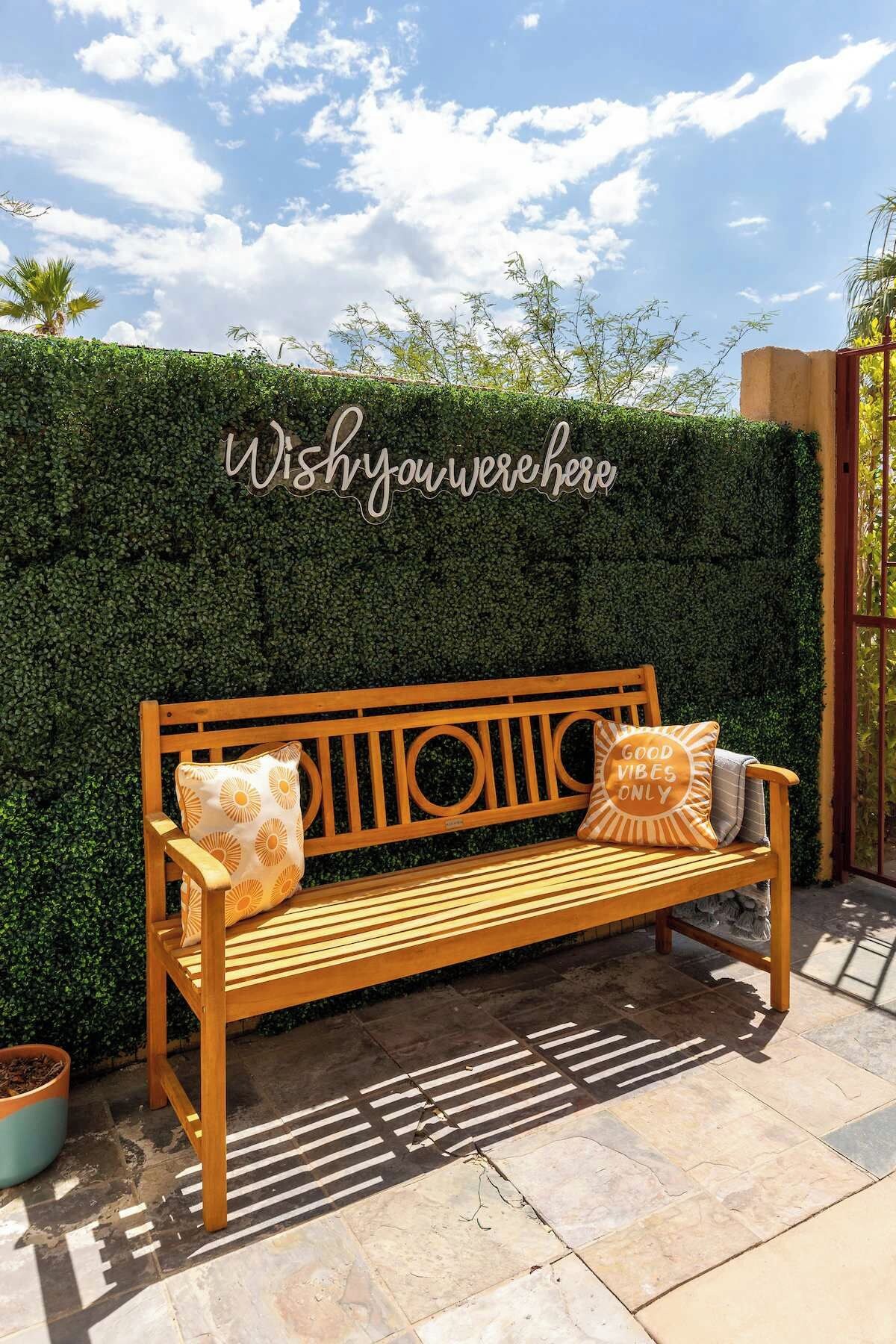 A wooden bench with cushions in front of a hedge with a sign that reads 'Wish you were here'.