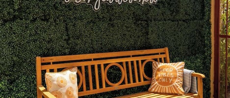 A wooden bench with cushions in front of a hedge with a sign that reads 'Wish you were here'.