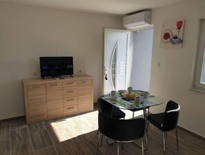 A small, well-lit room with a wooden cabinet, a TV, a glass table with chairs, and a door leading to a balcony.