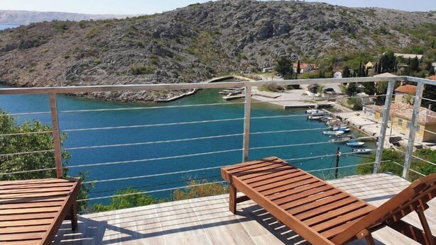 Terraza de madera con dos sillones de descanso con vista a un puerto deportivo con barcos, un promontorio rocoso y un pequeño pueblo al fondo.
