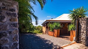 Una casa rustica con vialetto sterrato, portico in legno e piante tropicali.
