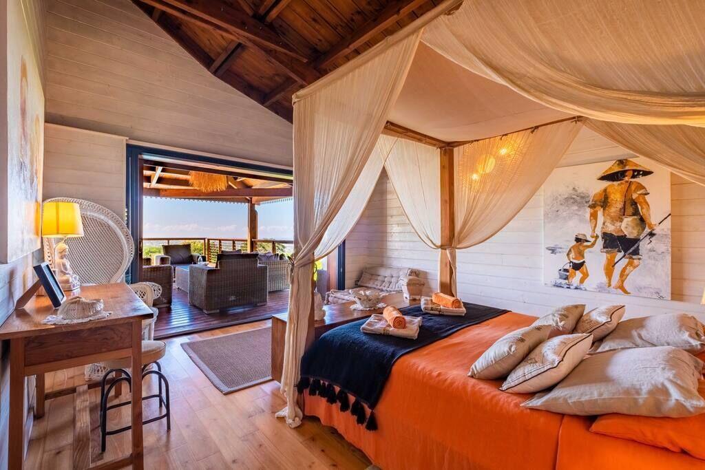 A room with a canopy bed, a desk with a lamp, and a view of the outside.