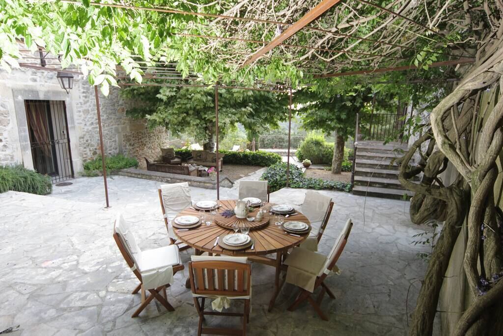 A round wooden table set for six with white chairs, surrounded by stone flooring, under a pergola with climbing plants.