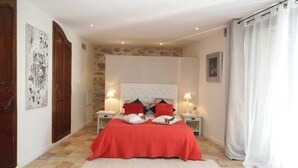 A bedroom with a large bed, two bedside tables, and a stone accent wall. - L'ESTAOU DE FINOU*** (Saint-Paul-et-Valmalle)