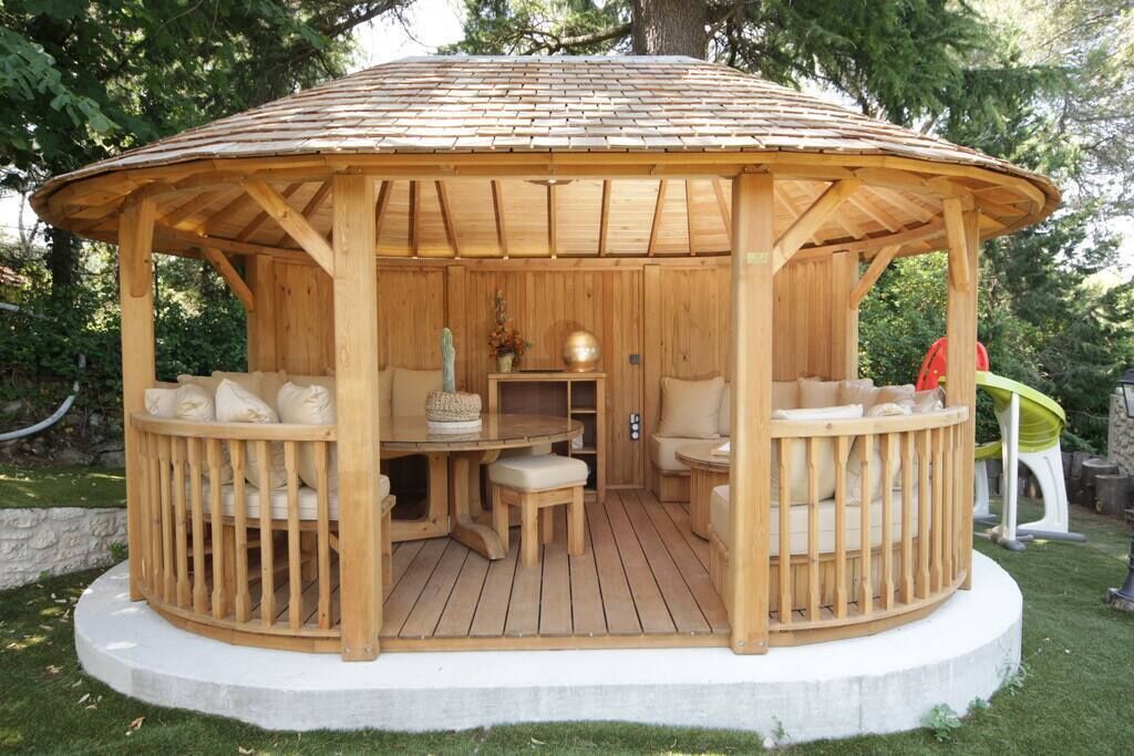 A wooden gazebo with a circular roof, a central table, and cushioned seating.