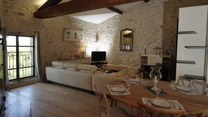 Un intérieur rustique avec des murs en pierre apparentes, un plafond en bois, un canapé blanc, une table à manger dressée pour quatre personnes et un téléviseur.