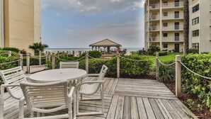 A wooden deck with a round table and chairs, overlooking a beach and a building with balconies. - Castle Reef - 121 Steps from the Sand (New Smyrna Beach)
