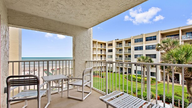 A balcony with white metal furniture, a view of the ocean, and a building with multiple balconies and greenery.