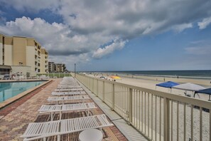 A poolside area with lounge chairs and umbrellas, a beach view, and a building with multiple floors.