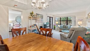 A spacious living room with a wooden dining table, comfortable seating, a television, and large windows offering a view of the outdoors.