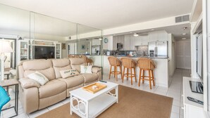 Ein modernes Wohnzimmer mit einem beigen Sofa, einem weißen Couchtisch und einer Küchenzeile mit weißen Schränken und einem Edelstahl-Kühlschrank.