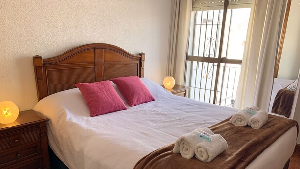 Un lit bien fait, avec une tête de lit en bois, deux oreillers, une couverture marron et des serviettes enroulées posées sur le lit.