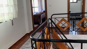 Une chambre d’hôtel avec un escalier en bois, un coin repas avec une table et des chaises, et une vue sur la chambre voisine, où l’on aperçoit un lit et des rideaux.