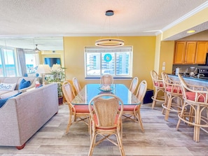 A modern living room with a sofa, a dining table with chairs, a kitchen area, and a large window with a view.