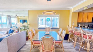 A spacious living area with a sofa, dining table, and chairs. There is a kitchen area with wooden cabinets and a window with a view.