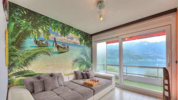 Un saló modern amb un mural gran que representa una platja tropical, un sofà gris amb coixins, un tauler d'escacs i un gran finestral corredís que dona a un balcó amb vistes a muntanyes i aigua.