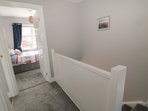A narrow hallway with a carpeted floor leading to a bedroom with a bed, a window with curtains, and a picture on the wall. - Fisherman's Row (Morpeth)