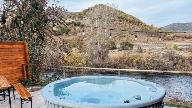 A hot tub with a wooden bench and a table, surrounded by a railing, overlooking a river and hilly landscape. - Charming 2 BR riverside stopover in Gold Hill. Private hot tub, river access, and just down the road from Del Rio Vineyards. Perfect base for exploring the Rogue Valley. Pet-friendly! (Gold Hill)