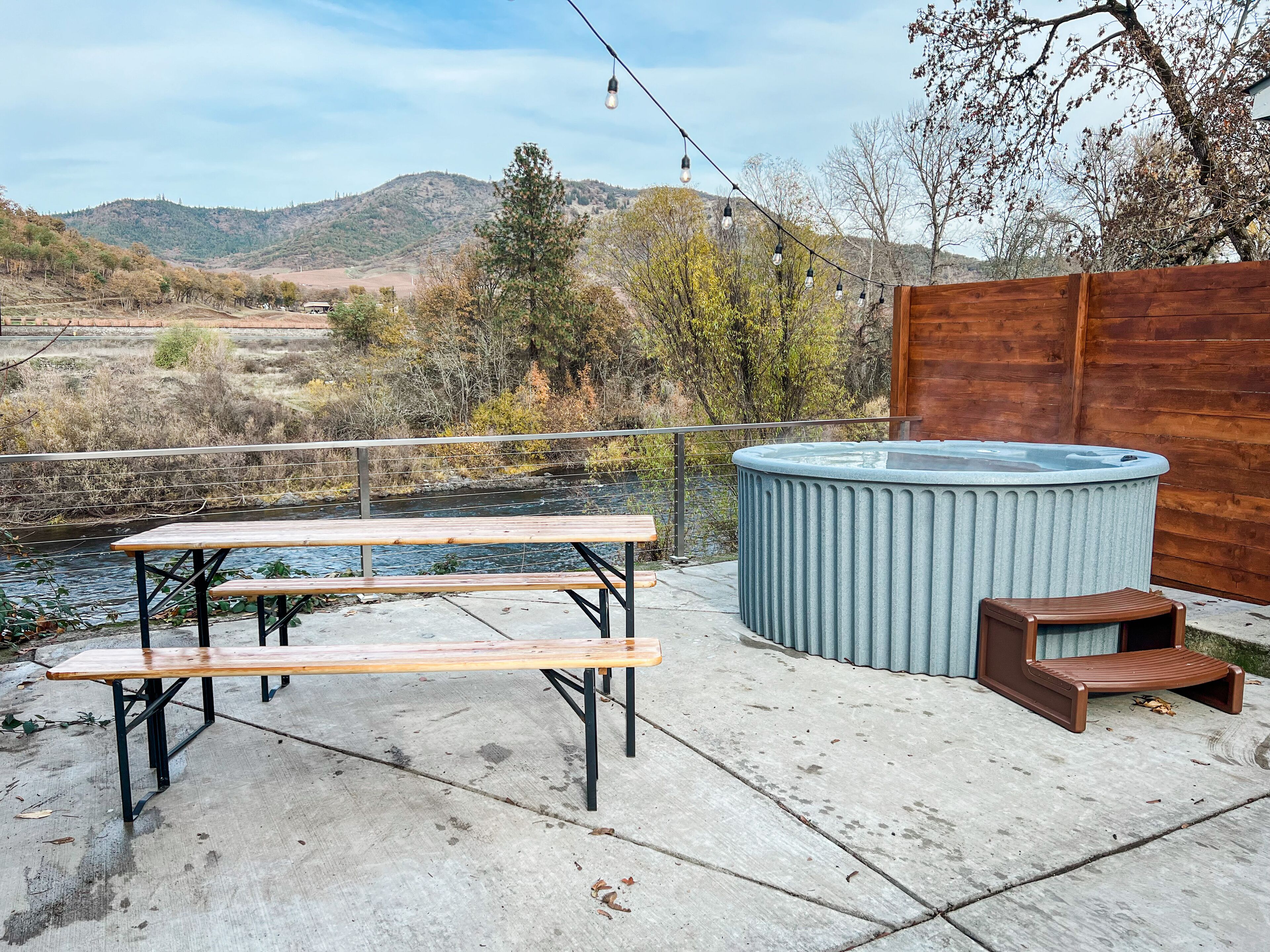 Eine Terrasse mit Whirlpool, Picknicktisch und Lichterkette mit Blick auf einen Fluss und Hügel.