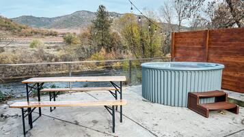 Eine Terrasse mit Whirlpool, Picknicktisch und Lichterkette mit Blick auf einen Fluss und Hügel.