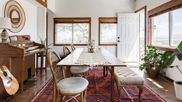 Un comedor con una larga mesa de madera, sillas, un piano y un tapete rojo estampado.