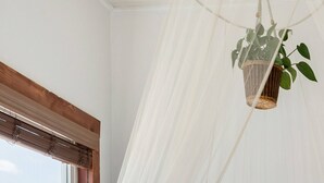 Una cama con un edredón blanco acolchado, dos almohadas y una manta azul. Un dosel blanco cuelga del techo, y una cesta tejida con una planta está colgada del dosel.