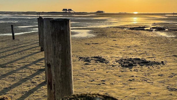 En trästolpe på en sandstrand med solen som går ner i horisonten i bakgrunden.