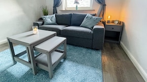 Ett modernt vardagsrum med en grå soffa, ett litet bord, en blå matta och ett fönster med gardiner.