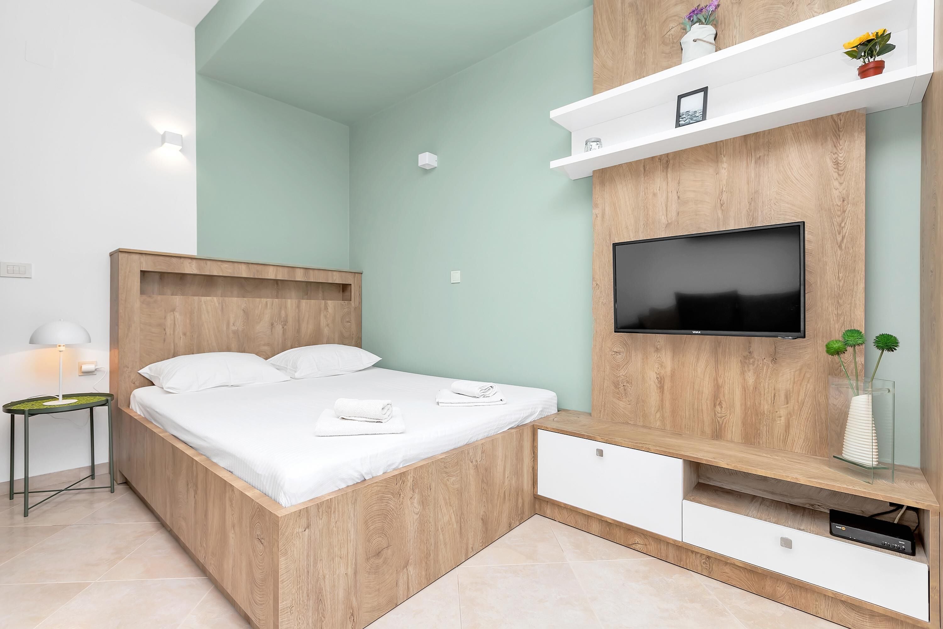 A modern bedroom with a wooden bed, a flat-screen TV mounted on a wooden wall, a small bedside table, and a white nightstand.