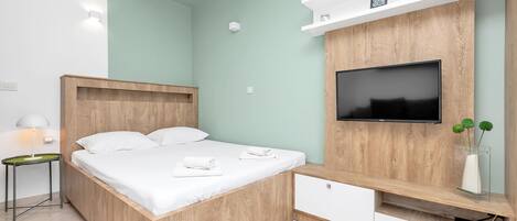 A modern bedroom with a wooden bed, a flat-screen TV mounted on a wooden wall, a small bedside table, and a white nightstand.