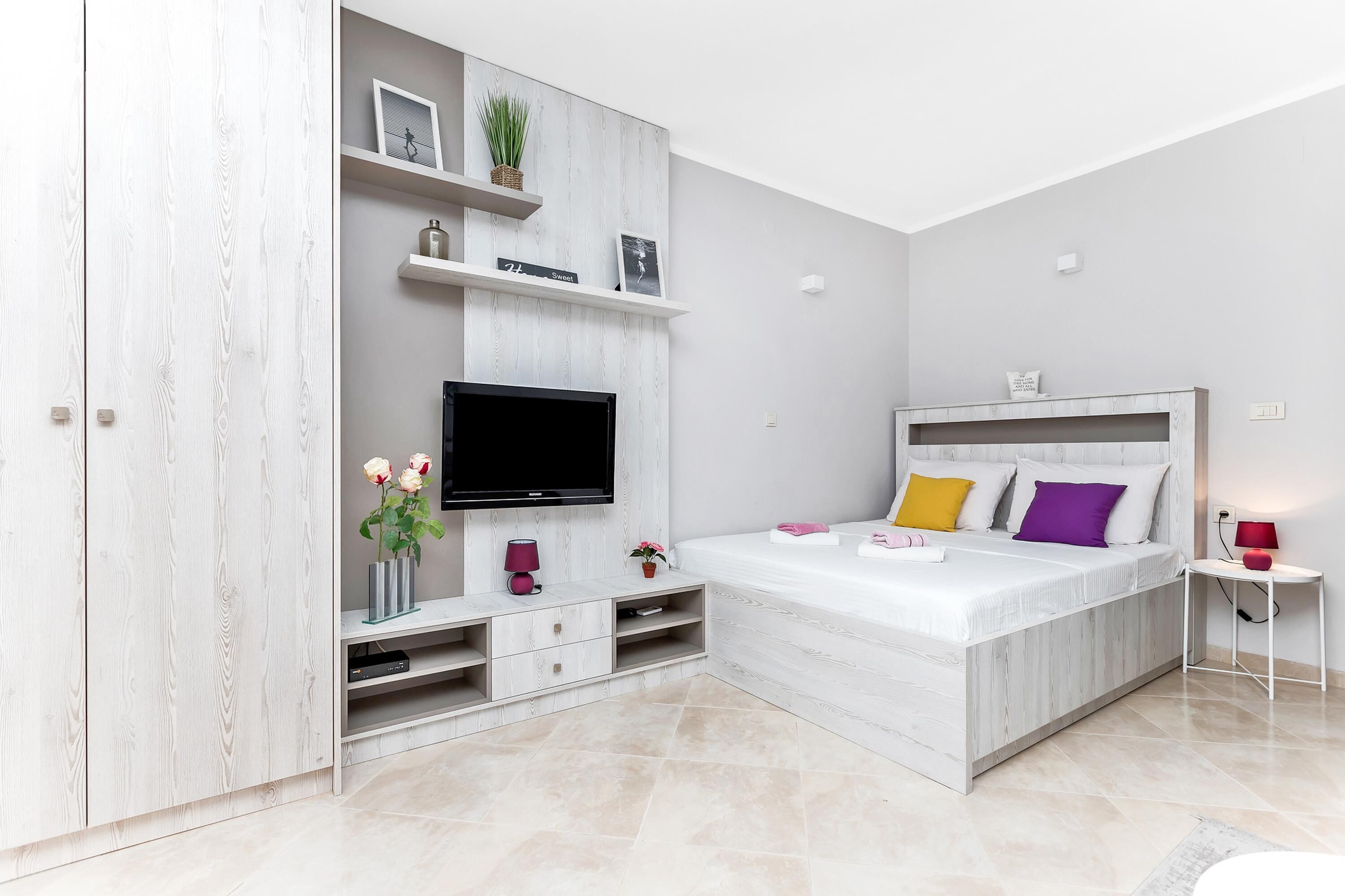 Un dormitorio moderno con televisión integrada, una cama con almohadones de colores, una mesita auxiliar pequeña y un armario blanco.