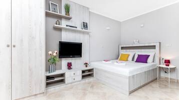 Un dormitorio moderno con televisión integrada, una cama con almohadones de colores, una mesita auxiliar pequeña y un armario blanco.