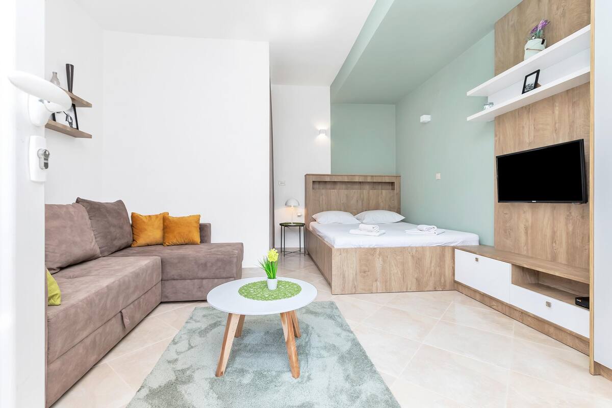 Una sala de estar moderna con un sofá, una mesa de centro redonda, una cama con cabecera de madera y un televisor fijado a la pared.