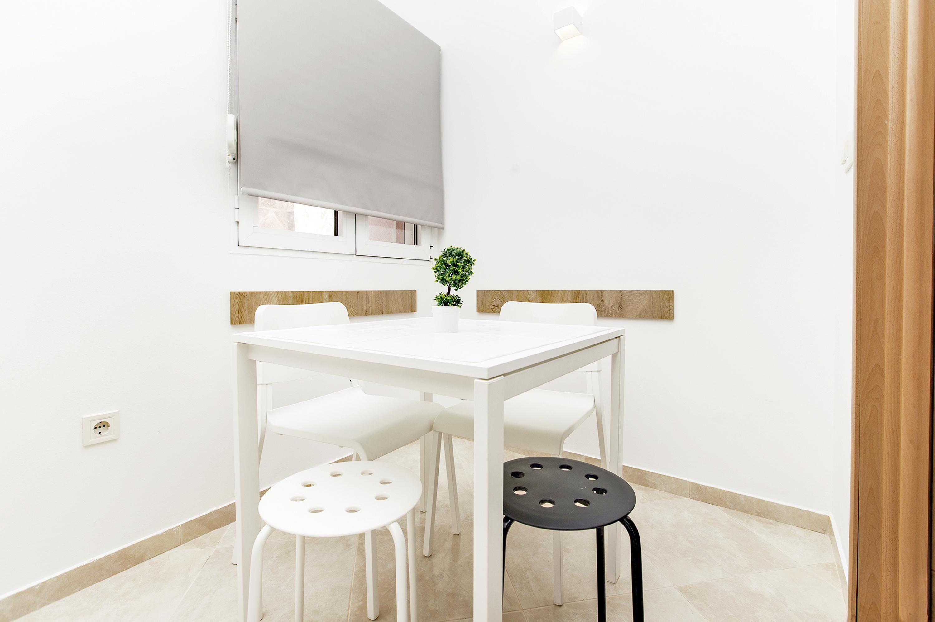 A small, white dining area with a table and four chairs, a potted plant, and a window with a blind.