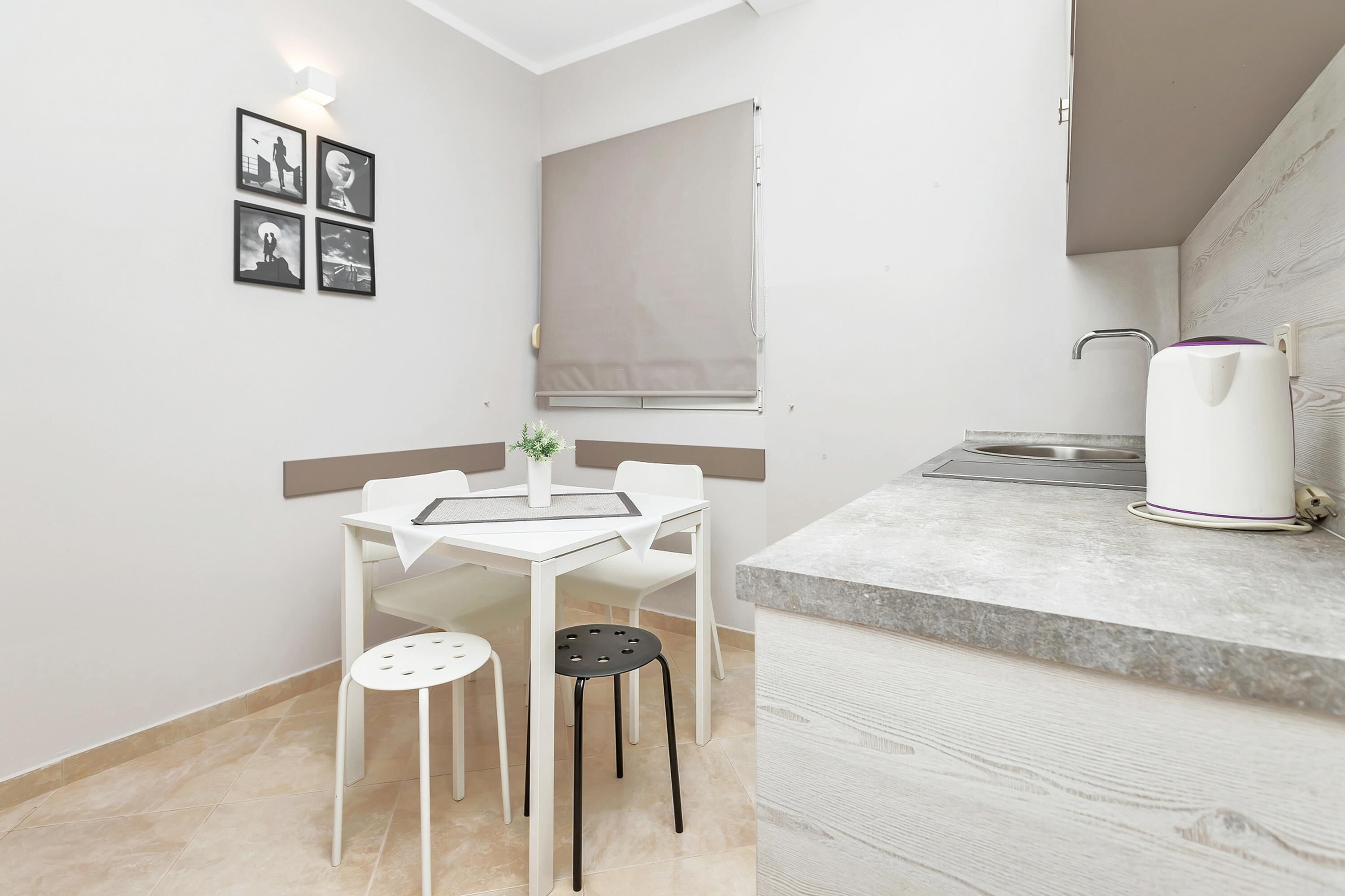 Una cocina pequeña y moderna con encimera blanca, un lavamanos y un hervidor. Hay una zona de comedor con una mesa y sillas, y una ventana con persiana.