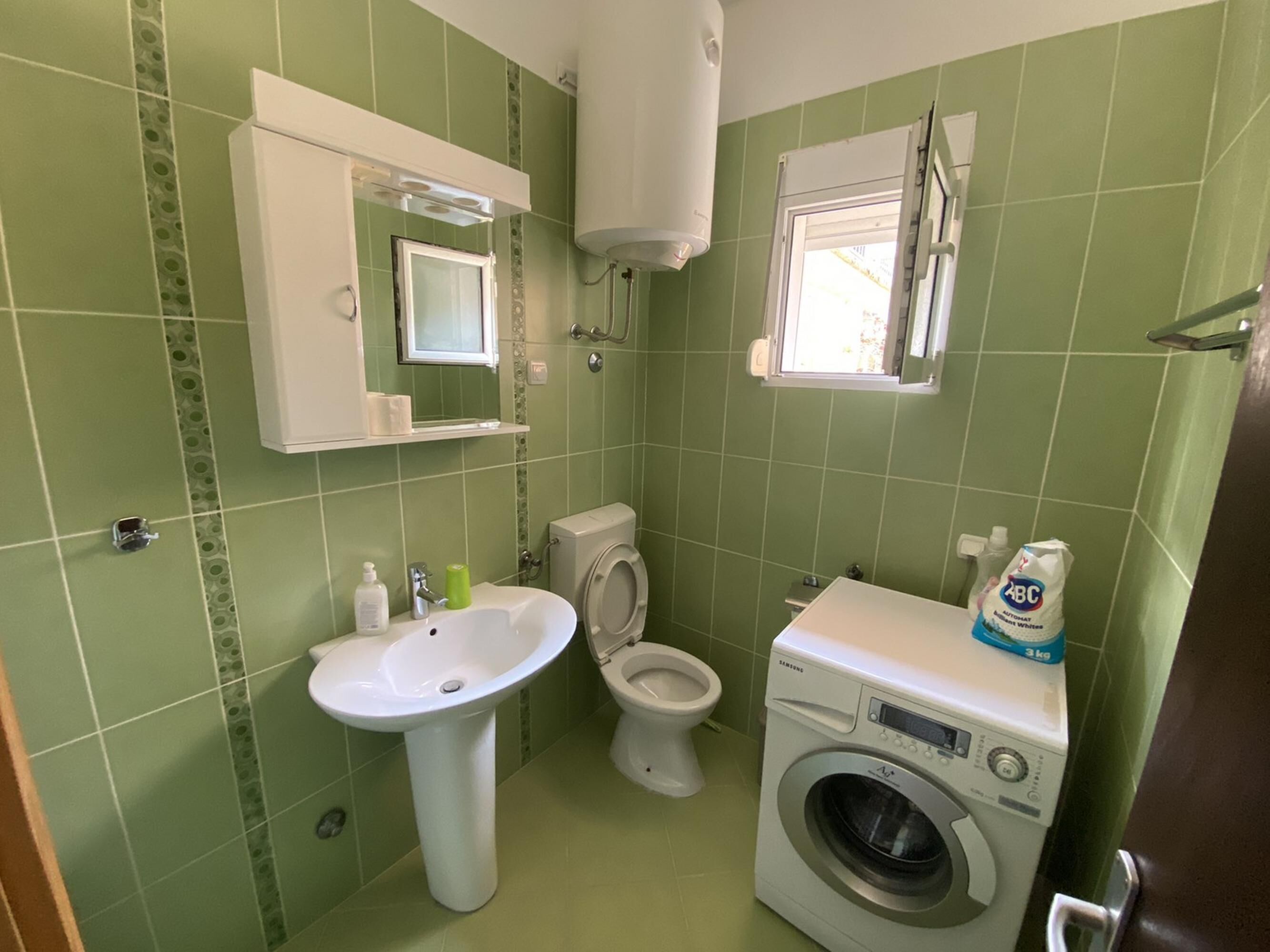A small bathroom with green tiles, a white sink, a toilet, and a washing machine.