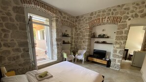 Ein Schlafzimmer mit Steinwänden, einem Bett mit weißen Leinentüchern, einem Fernseher, einem Stuhl und einem kleinen Tisch.