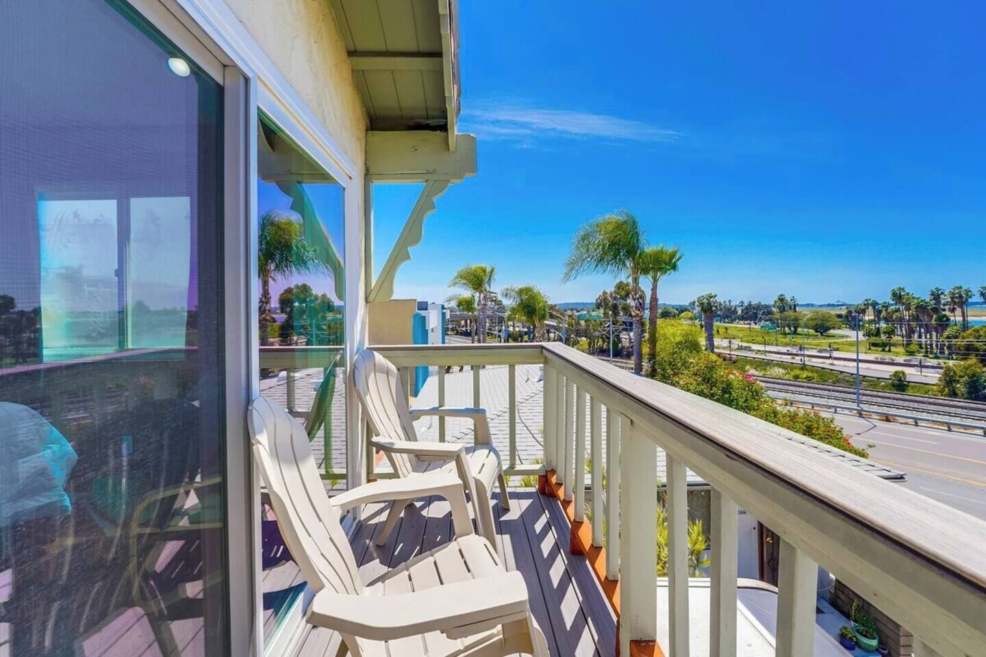 A balcony with chairs, a railing, and a view of palm trees and a road.