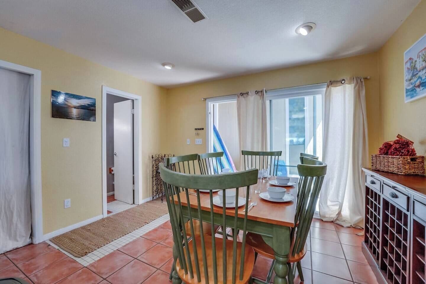 Un comedor con una mesa dispuesta para cuatro personas, sillas, una ventana con cortinas y vistas al exterior.