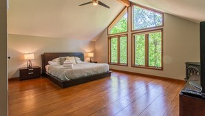 Un dormitorio con una cama grande, dos mesitas de noche con lámparas, un ventilador de techo y un amplio ventanal con vistas a árboles.