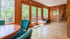 Una cabaña de madera con ventanas amplias, una zona de comedor con una mesa redonda y sillas azul turquesa, y una cocina con una puerta de madera.