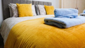 Een net opgemaakt bed met een grijs hoofdeinde, een gele deken en twee blauwe handdoeken.
