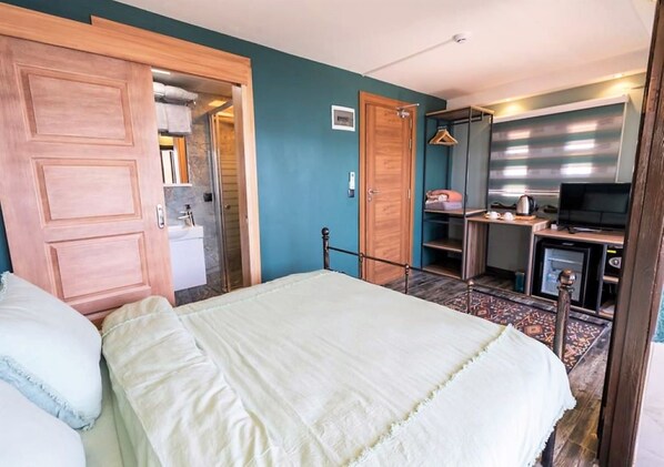 A bedroom with a bed, a wooden wardrobe, a desk with a television, and a bathroom visible through an open door.