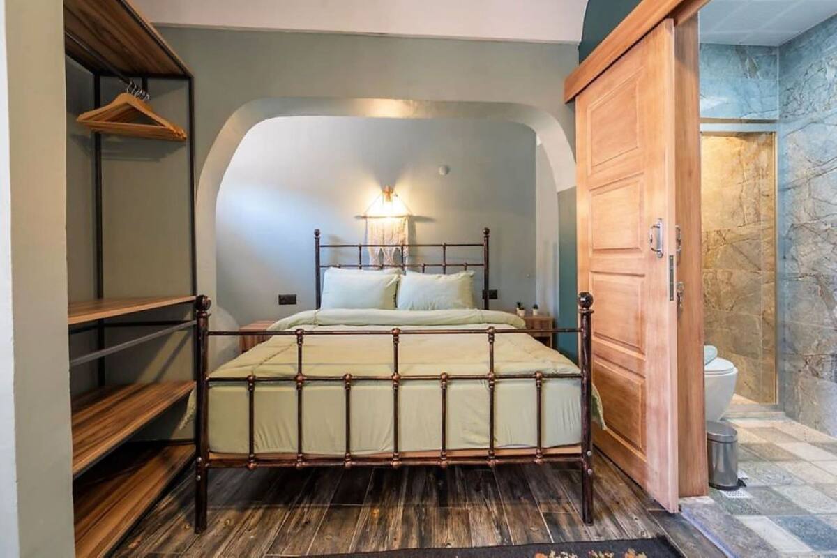 A bedroom with a bed, wooden furniture, a hanging rack, and a bathroom with a toilet and shower.