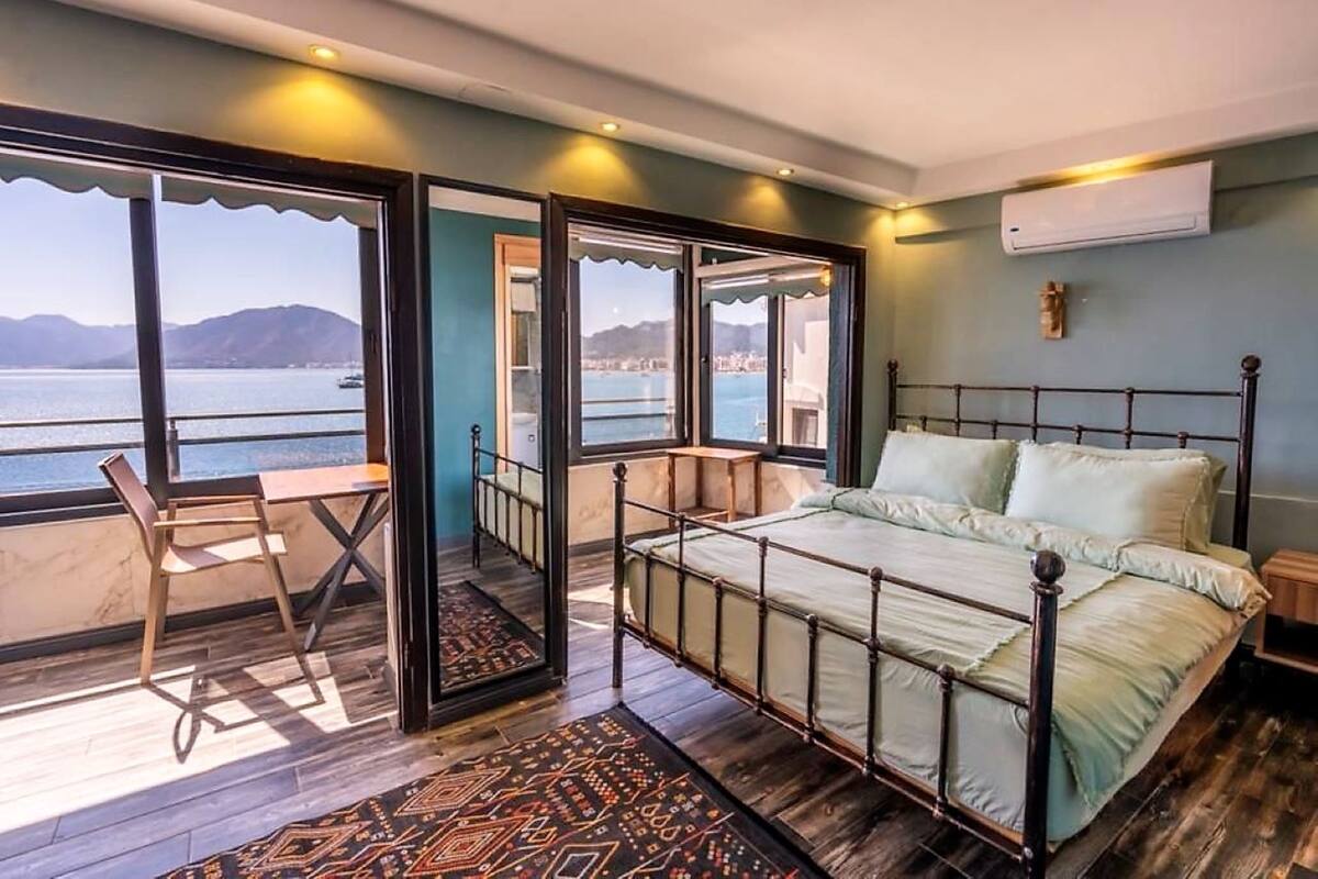 A bedroom with a bed, a chair, a small table, and a view of the sea.