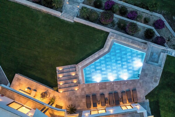 An aerial view of a swimming pool with a geometric design, surrounded by a stone deck and a small patio area with seating.