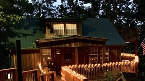 Uma cabana de madeira com varanda decorada com luzes de cordão, um pequeno deck e um caminho de pedras que leva à entrada.