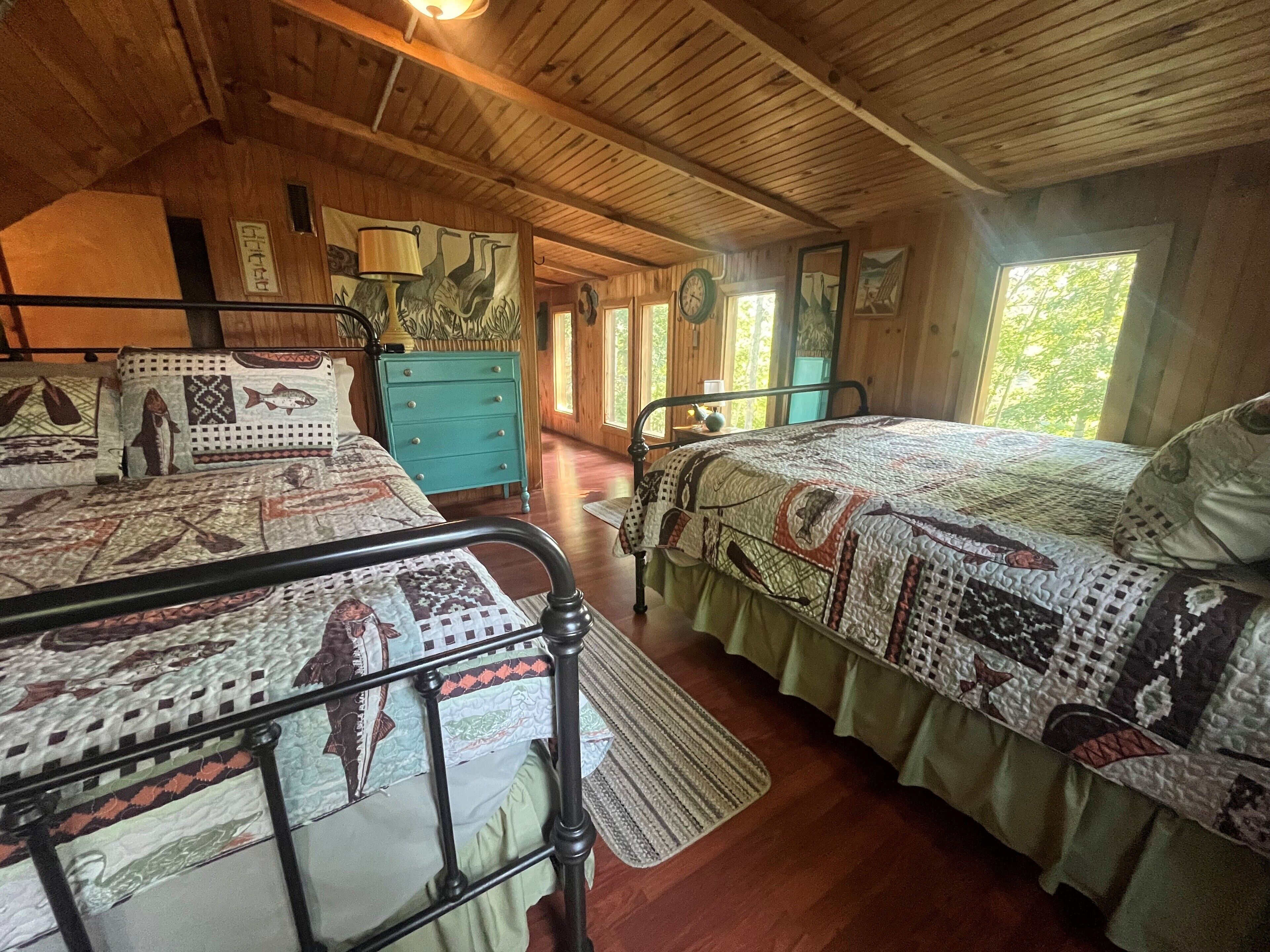 Ein gemütliches Blockhauszimmer mit zwei Betten, einer Holzbalkendecke, einer Uhr und einem Fenster mit Blick auf Bäume.