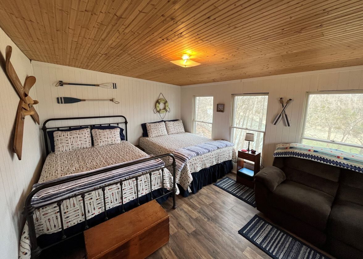 A cozy cabin-style bedroom with two beds, a wooden ceiling, and a large window.