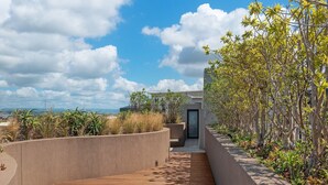 Un chemin moderne aménagé à l’extérieur, avec un revêtement de sol en bois, bordé de petits murets en béton et d’une végétation luxuriante, qui mène à un bâtiment doté d’une porte en verre.